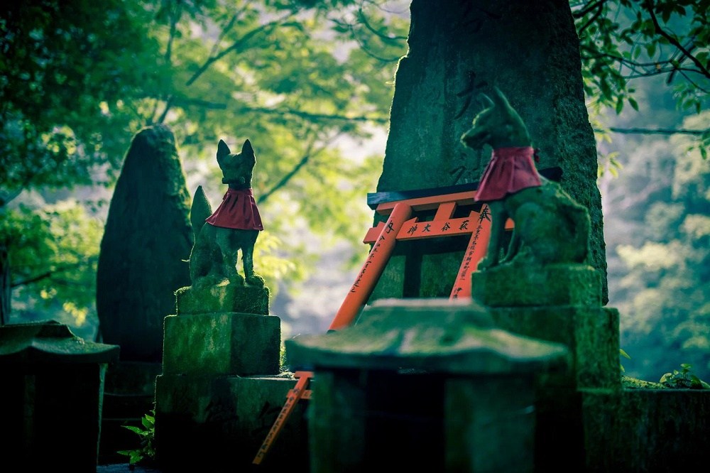 Statues or monuments in a wooded area, trees nearby, nearest statue is of a fox next to a cloaked human.