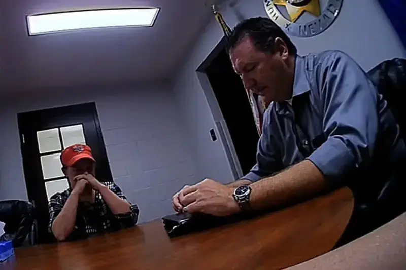 A woman wearing a baseball cap sitting at a table across from a male investigator in an office.