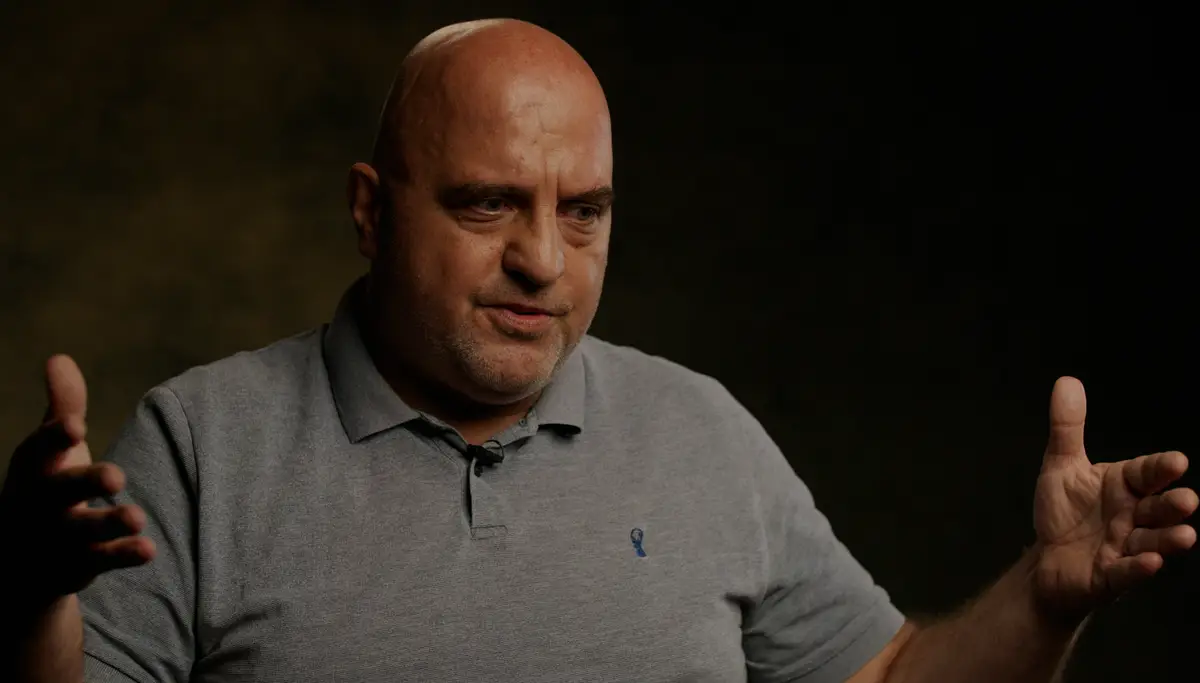 A close-up portrait of Andrew Chaggar, a bald man with light skin, speaking during an interview in front of a dark, blurred background.